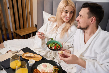 Young blonde woman and her husband are having breakfast in hotel bedroomの写真素材