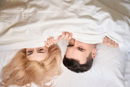 Happy newlyweds nestled comfortably under a blanket on large bed