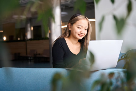 Smiling asian female IT employee watching laptop on sofa in coworking office
