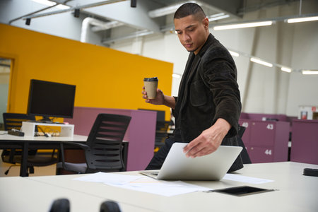Serious guy standing while holding glass and looking at laptop