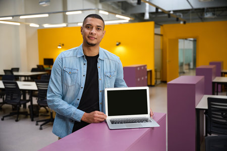Happy young employee standing in office while showing laptop