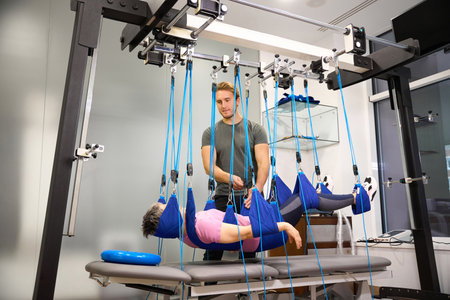 Young male trainer helping middle aged woman hanging on suspension medical ropes