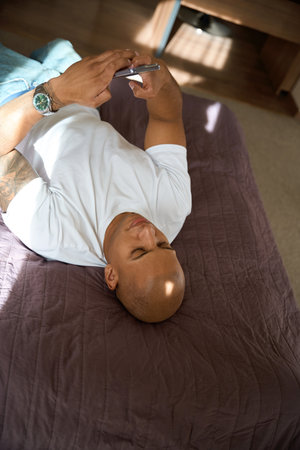Focused african american man lying on bed and using smartphone in hotel room