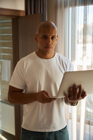 Serious african american man with laptop looking at camera in hotel room at dayの写真素材