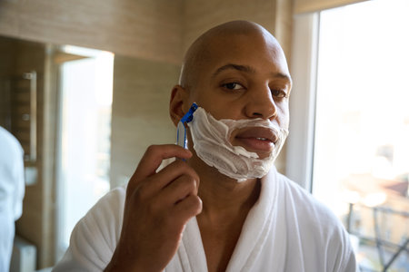 Serious young man shaving with razor and looking at camera in bathroomの写真素材