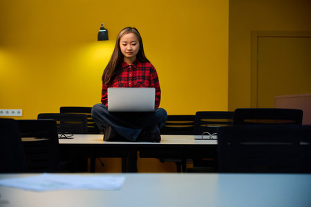 Front view of focused asian female IT employee using laptop sitting on desk