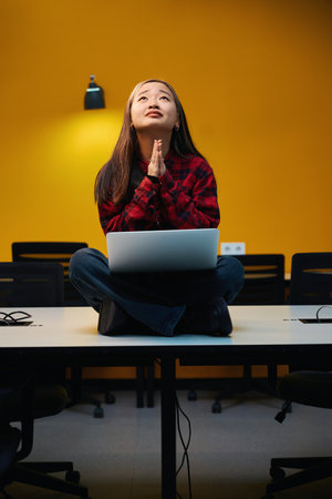 Young tense asian female IT employee with praying hands using laptop on desk