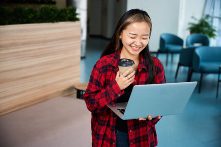Laughing asian female IT employee watching laptop in hall of coworking officeの写真素材