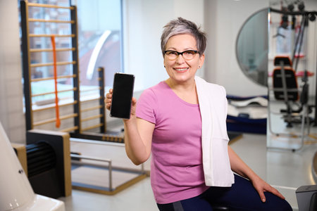 Adult smiling caucasian woman showing smartphone and looking at camera in gym