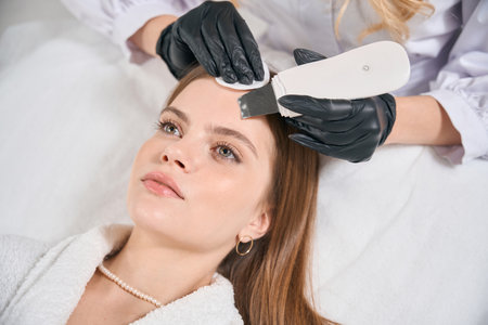 Beautician doing ultrasonic cleaning of face skin of young beautiful woman