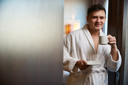 Pleased man with beverage standing in hotel room doorwayの写真素材