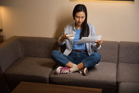 Focused woman looking through documents sitting on sofaの写真素材