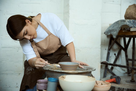 Focused female artist in apron making clay pottery on a spin wheel in creative studio