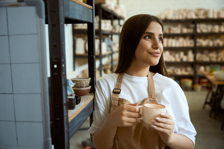 Female ceramics studio owner in apron with cup posing in workshop