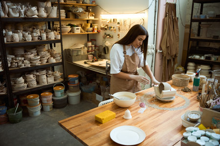 Pretty female ceramist in apron taking a clay mug out of the mold in studio