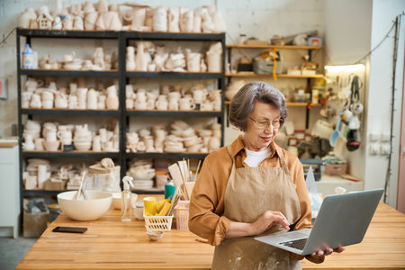 Elderly woman stands in pottery room with laptop in her handsの写真素材