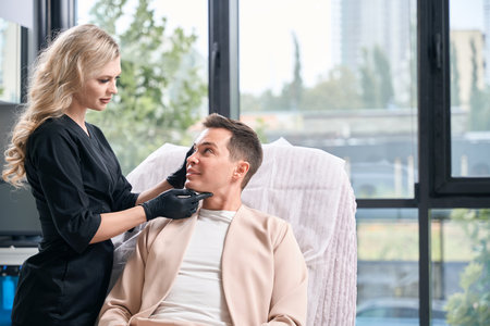 Woman examining face skin of adult male client, esthetic male face cleansingの写真素材