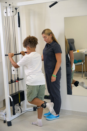 Young patient practices strength exercises on resistance equipment with therapists supervision