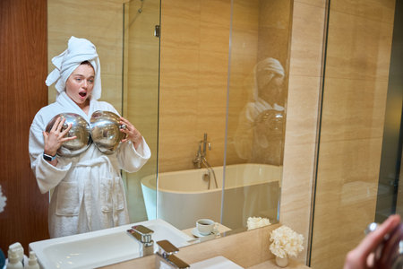 Surprised young woman in bathrobe holding metal bowls indoors