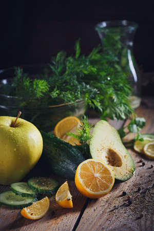 Background with assorted green vegetables: apple, avocado, cucumber, lemon and detox water on rustic wooden table top. Healthy food, detox, dieting, vegetarian and weight loss concept with copy space.の写真素材