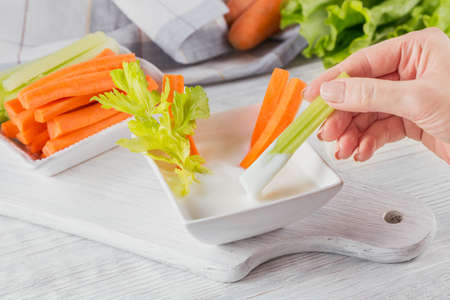 vegetable sticks. Fresh celery and carrot with yogurt sauce. Woman's Hand holding celery stick. Healthy and diet food concept.の写真素材