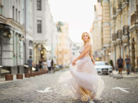 Pretty smiling young woman with long blond hair in elegant flying light dress running along the streetの写真素材