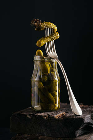 Gherkins, pickled cucumber on a fork, bowl of marinated vegetables on dark wooden background. Clean...