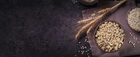 Bowl of cooked peeled barley grains porridge with ears of wheat on dark wooden background. Cooking...
