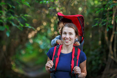 Mother with toddler child in backpack carrier is hiking in forest. Tourist is carrying a baby on his...