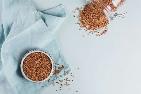 Organic uncooked scattered buckwheat grain in a bowl and glass jar on light blue background. Healthy and diet food concept. top view, flat layの写真素材