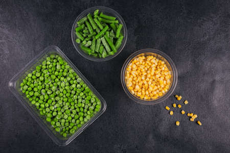 Plastic container with different organic deep frozen vegetables on black table. Green peas, sweet...