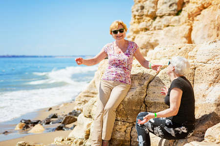 Two elderly women are happy to meet each other, sitting on rock on the seashoreの写真素材