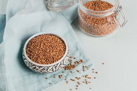 Bowl of dry raw buckwheat groats and glass jar on a light blue background. Cooking buckwheat...