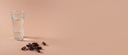 Panoramic wide banner with diet, nutrition, healthy eating concept. Oil filled capsules of food supplements and glass of water for pouring brown softgels capsules on beige backgroundの写真素材