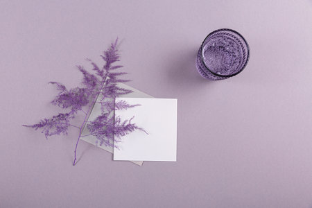 Blank greeting card mockup with envelope, white empty paper, glass of sparkling water and dried plant for Mother's or women's Day. Free space for message or design on pastel violet backgroundの写真素材