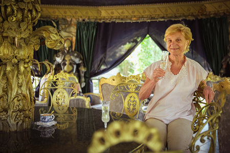 Mature attractive woman traveler sitting alone on the terrace of coffee shop in beautiful park in Bulgaria and drinking champagne. Active life of the elderly in retirement, active seniorsの写真素材