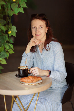 Woman sitting at table and enjoying cup of coffee and croissant in cafeの写真素材