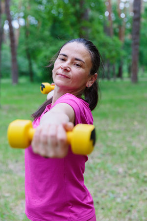 Mature Woman Raising Arms with Dumbbells in Parkの写真素材