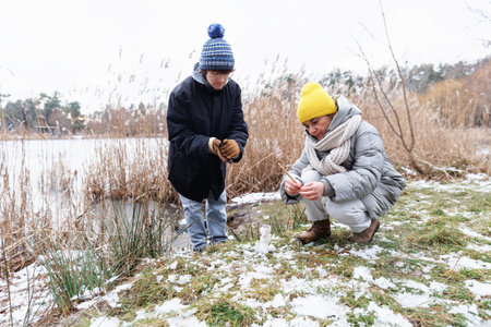 Mother and teenage son building mini snowman by lake in winterの写真素材