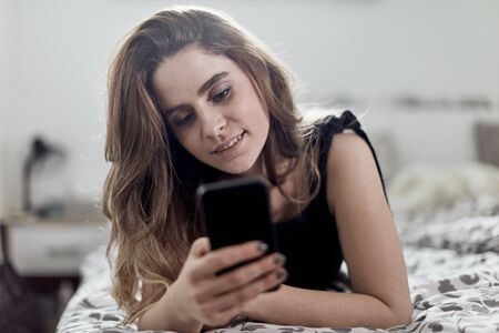 young brunette looking at her smart phone while laying on cosy bedの写真素材