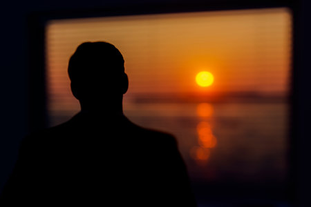 A man stands in a room and looks out the window at the setting sun.の写真素材