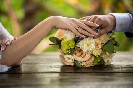 Hands of the bride and groom on a wedding bouquet. Close-up. Wedding rings in focus. Bouquet on a wooden table. Horizontal frame.の写真素材