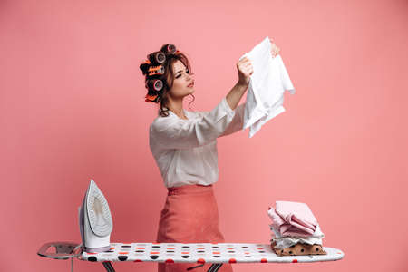 A pretty housewife irons clothes, carefully examines ironed clothes. Isolated on pink background.の写真素材