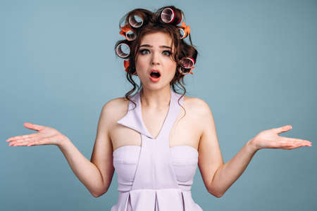 Surprised housewife spread her arms against the blue wall. The girl with curlers on her hair and in a dress is shocked.の写真素材
