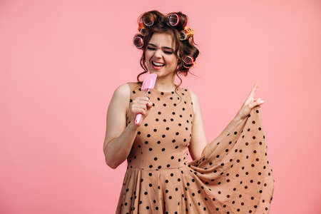 Smiling, joyful girl in a polka dot dress, beating eggs, cooking on the background of a rope with clothes. Housewife with curlers on her hair on a pink background.の写真素材