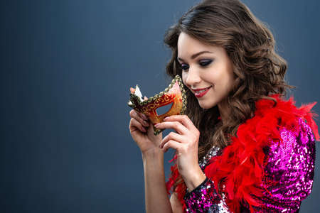 Smiling woman is holding a carnival mask in a glittering dress and a boa around her neck.の写真素材