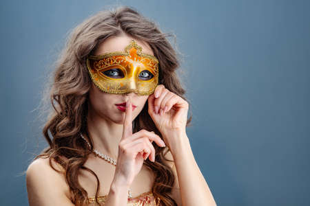Woman in a golden dress and carnival mask on a blue background touching her finger to her lipsの写真素材