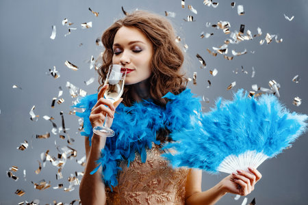 Woman in a blue boa is standing in the studio holding a fan of feathers in her hand and drinking...