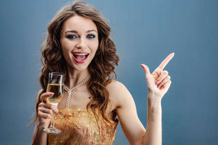 Brunette woman in golden dress and pearl necklace with raised glass of champagne celebratesの写真素材