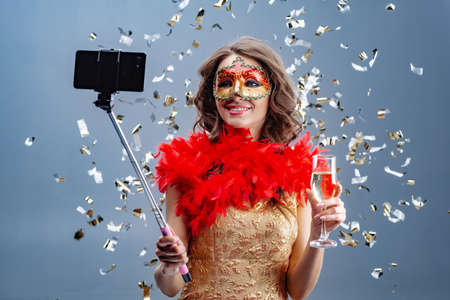 Photo of a smiling woman in  carnival mask and  gold dress with  raised glass makes  selfie on a mobile phone. Background with tinselの写真素材